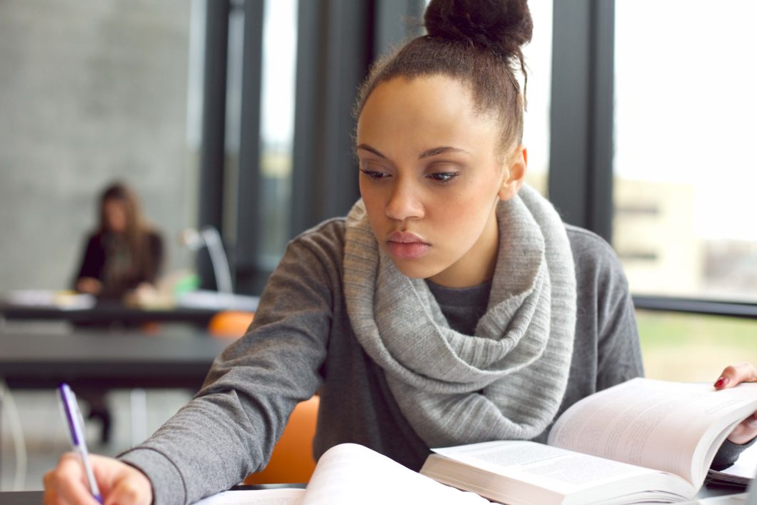 Close up image of a young female student doing assignments in library. Afro american woman taking notes from textbooks.