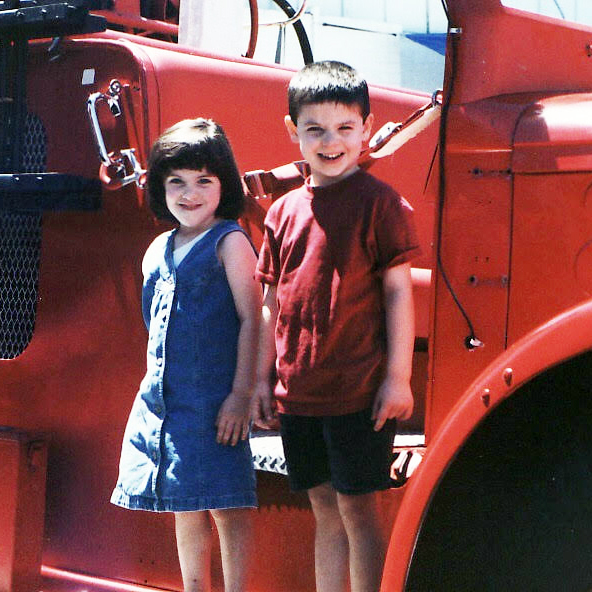 Rebecca Delevati and her twin brother, Stephen, as children standing in front of a fire truck.